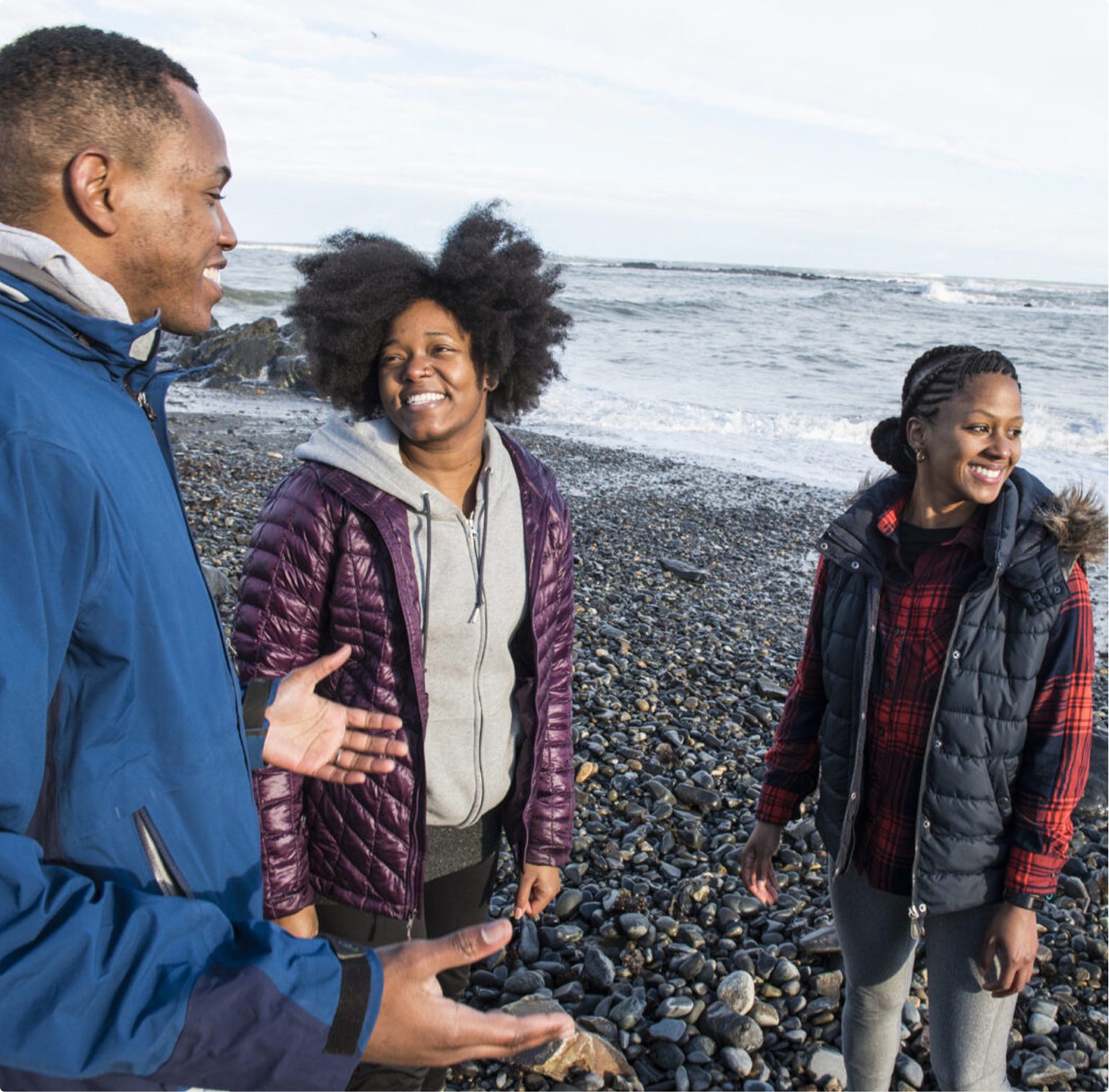 Three people in winter jackets on a rocky beach, laughing and smiling.