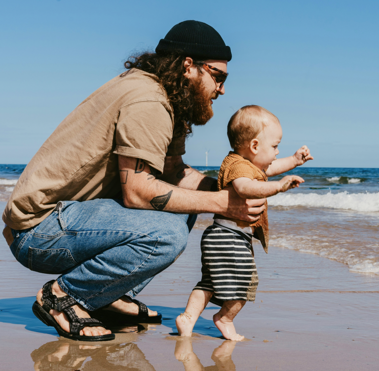 A man with long hair, a tan shirt, and jeans on a beach holding a baby.