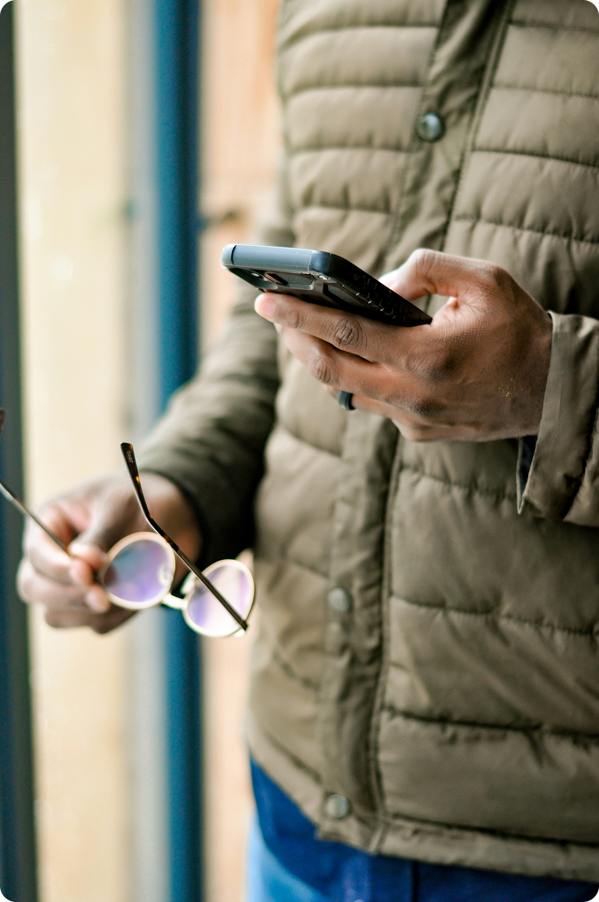 Person holding a phone in one hand and glasses in the other while wearing an olive green jacket.