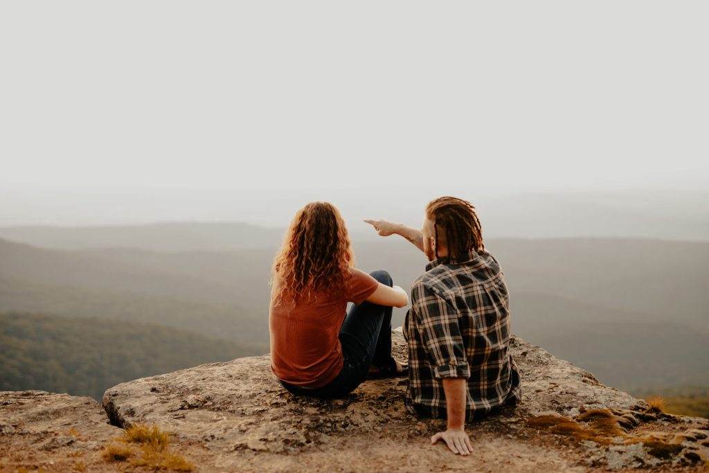 Two people sitting on rocks looking over the view. One person is wearing a plaid shirt and pointing in the distance.